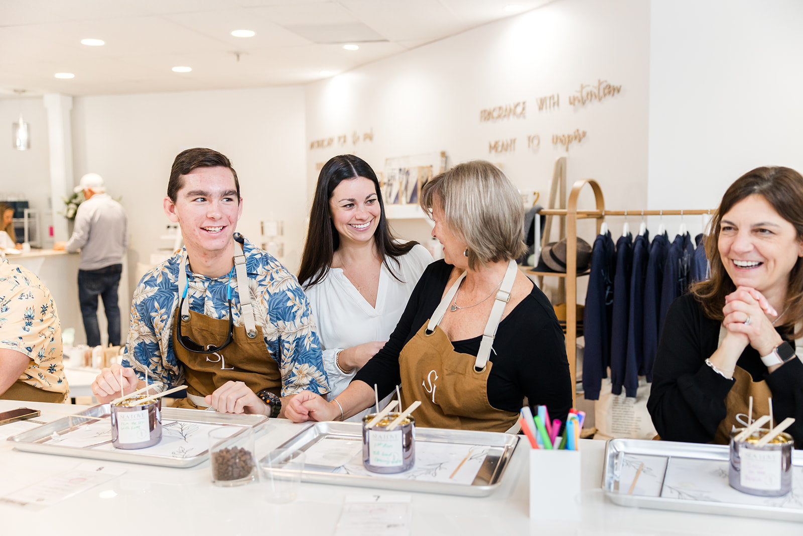 A group of joyful people wearing aprons participating in a fragrance workshop, interacting with each other around a table with candle making materials.