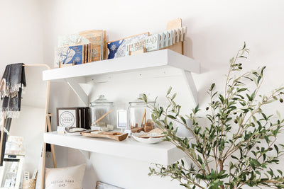 A serene corner with a white shelf displaying books, decorative jars, and wooden kitchenware, complemented by a green potted plant, against a clean, light-colored wall.