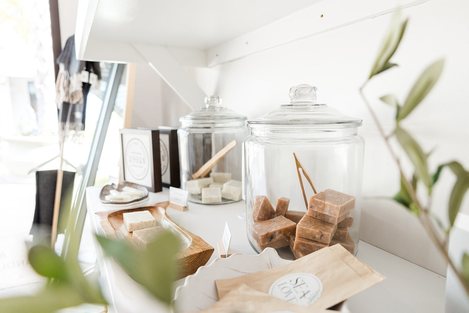 A serene and stylish shelf displaying an array of self-care products, including two glass jars—one open with wooden tongs inside—and eco-friendly soaps, all bathed in soft, natural light.