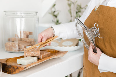 A person in a brown apron and white shirt presenting a selection of artisanal soaps arranged on a wooden tray, with a clear glass jar lid in hand and bathroom decor in the background.
