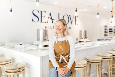 A cheerful women proudly sits in her bright and modern candle bar, embodying the fresh and coastal vibe of the establishment.