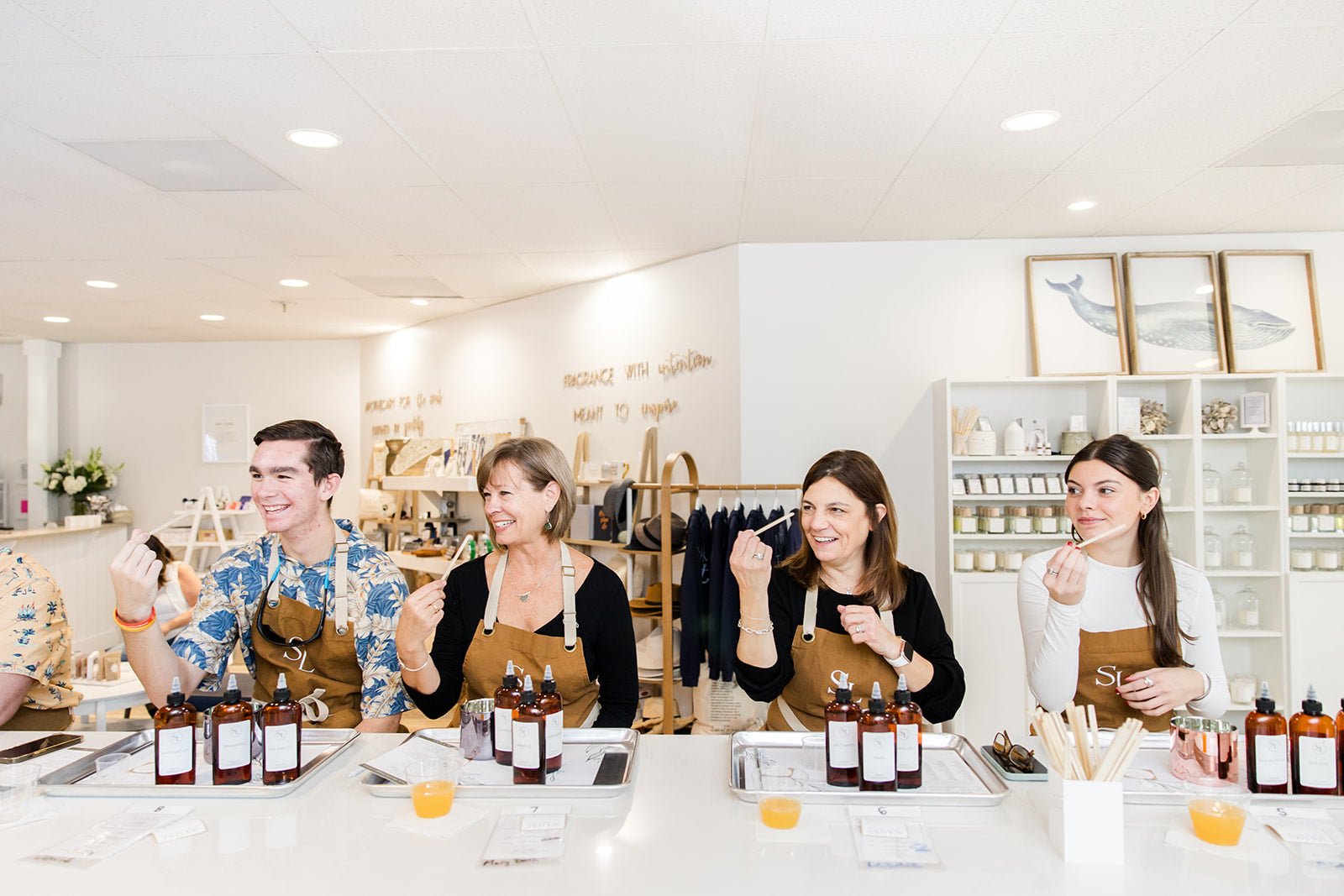 A group of people joyfully engaging in a fragrance blending custom candle experience, with clear glass bottles and mixing tools on the white tables in a bright and modern studio space.