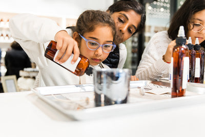 A young girl with glasses, closely observed by an adult, is carefully pouring fragrance oils from a brown bottle into a vessel, while another individual works alongside them.