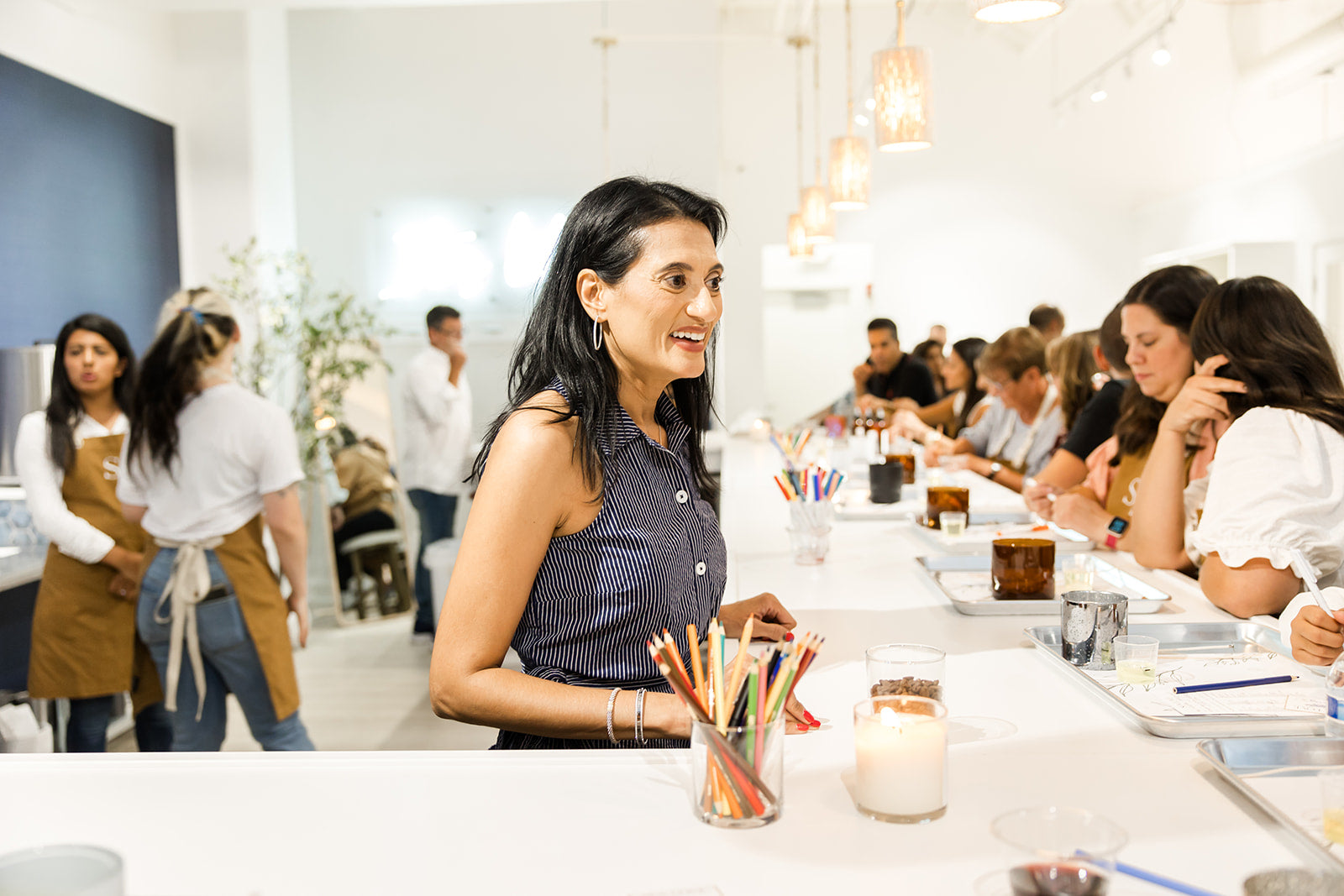 A smiling woman stands at the end of a long table filled with people engaged in a workshop or event in a bright, modern space.
