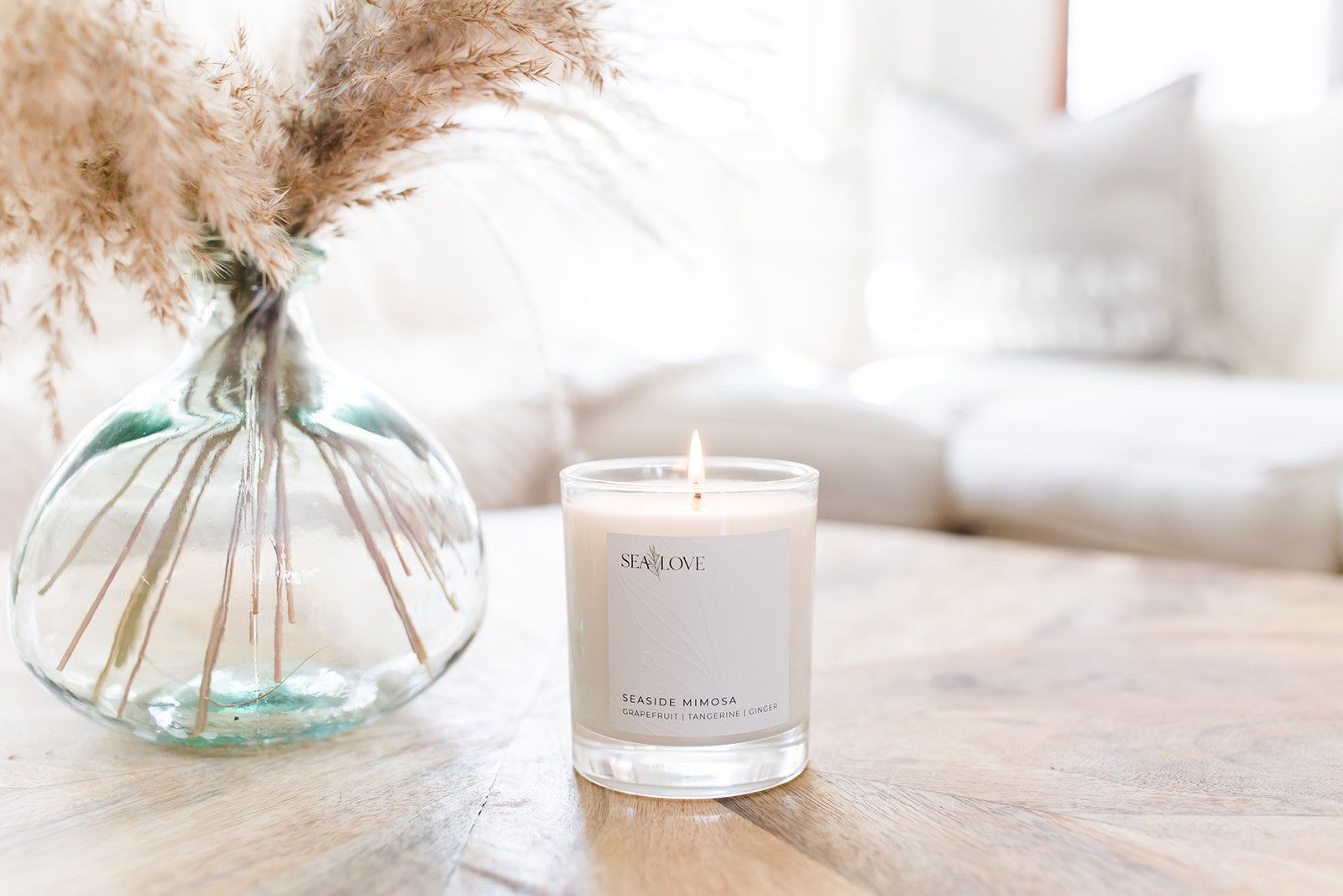 A serene setting with a lit scented candle named 'seaside mimosa' on a wooden table, accompanied by a vase of delicate pampas grass, conveying a warm and tranquil atmosphere.